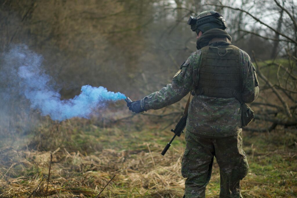 A soldier in camouflage uses a blue smoke signal during a tactical exercise in a forest setting.